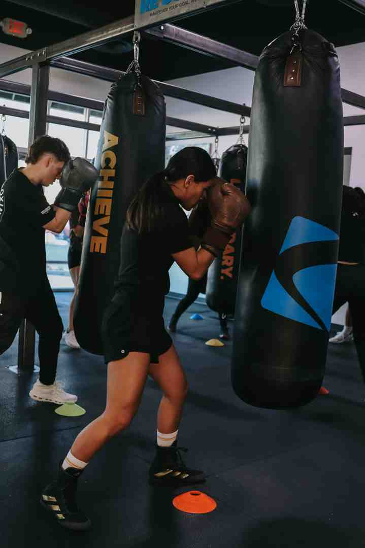 A woman in black shorts and cap drives both gloves deep into a Legends Boxing "ACHIEVE" heavy bag in a wide driving punch, while a male member behind her lands a cross on his own bag — cones on the floor, bags hanging overhead, the energy of a real class round in full motion