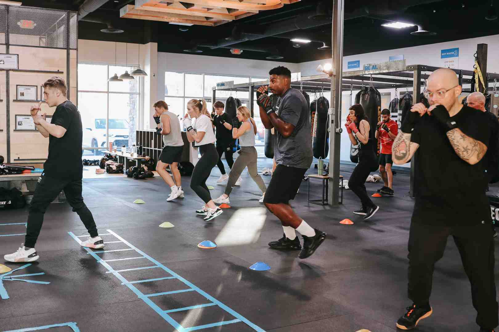 Wide-angle shot of a Legends Boxing class in full swing: eight or more members across age groups hold boxing guard stance on a gym floor mapped with an agility ladder and colored cones, a coach in gray leading the group, heavy bags hanging in the background — exactly what you'll find at any Legends Boxing location near you