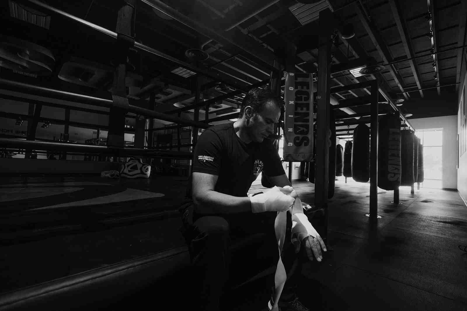 Black and white photograph: a man in a Legends Boxing t-shirt sits ringside on the edge of a boxing ring, head bowed in concentration as he carefully wraps white hand wraps around his right hand, rows of Legends Boxing heavy bags hanging in the dramatically lit background — the ritual of preparation before every session