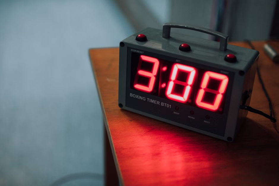 A Boxing Timer BT01 sits on a wooden surface, its red LED display showing "3:00" — the standard 3-minute round used in boxing training — casting a red glow on the wood beneath it, two more buttons visible on top for start and reset. Set yours before every session