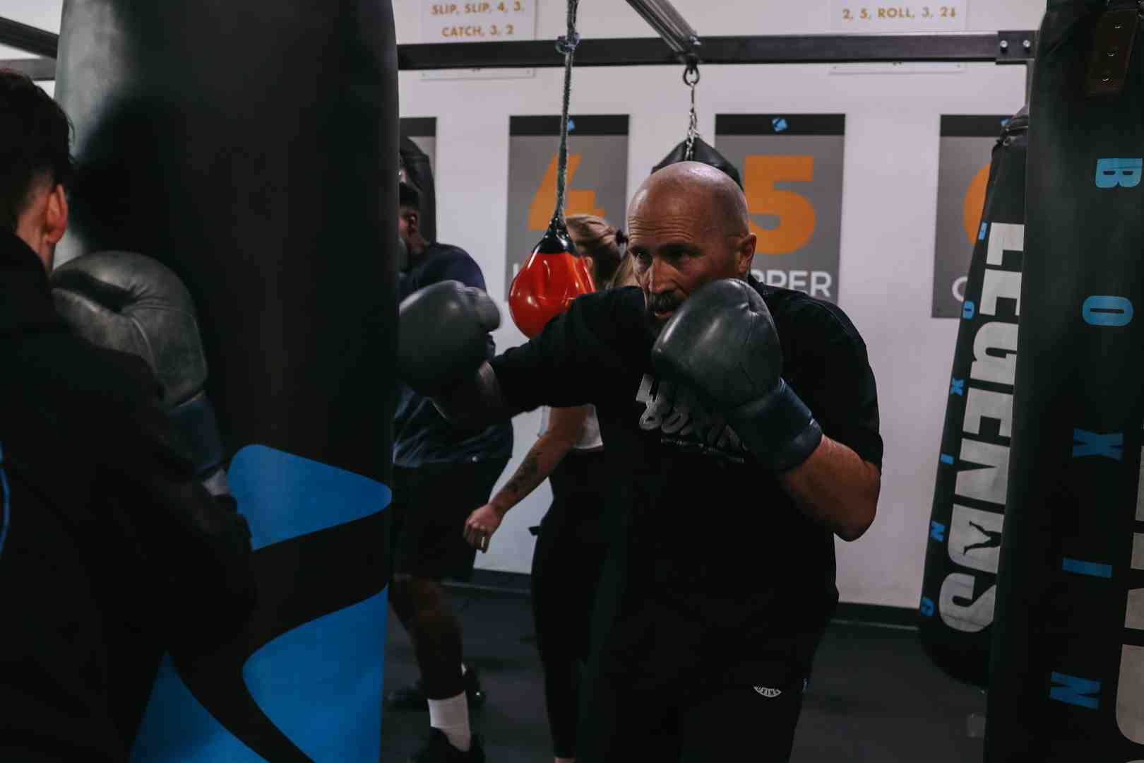 Bald Legends Boxing coach in black throwing a cross at a heavy bag during a busy group class — red speed ball and other members working in background, boxing combination board visible on wall ("SLIP, SLIP, 4, 3 / CATCH, 3, 2") — capturing the real-gym energy of a structured bag workout session
