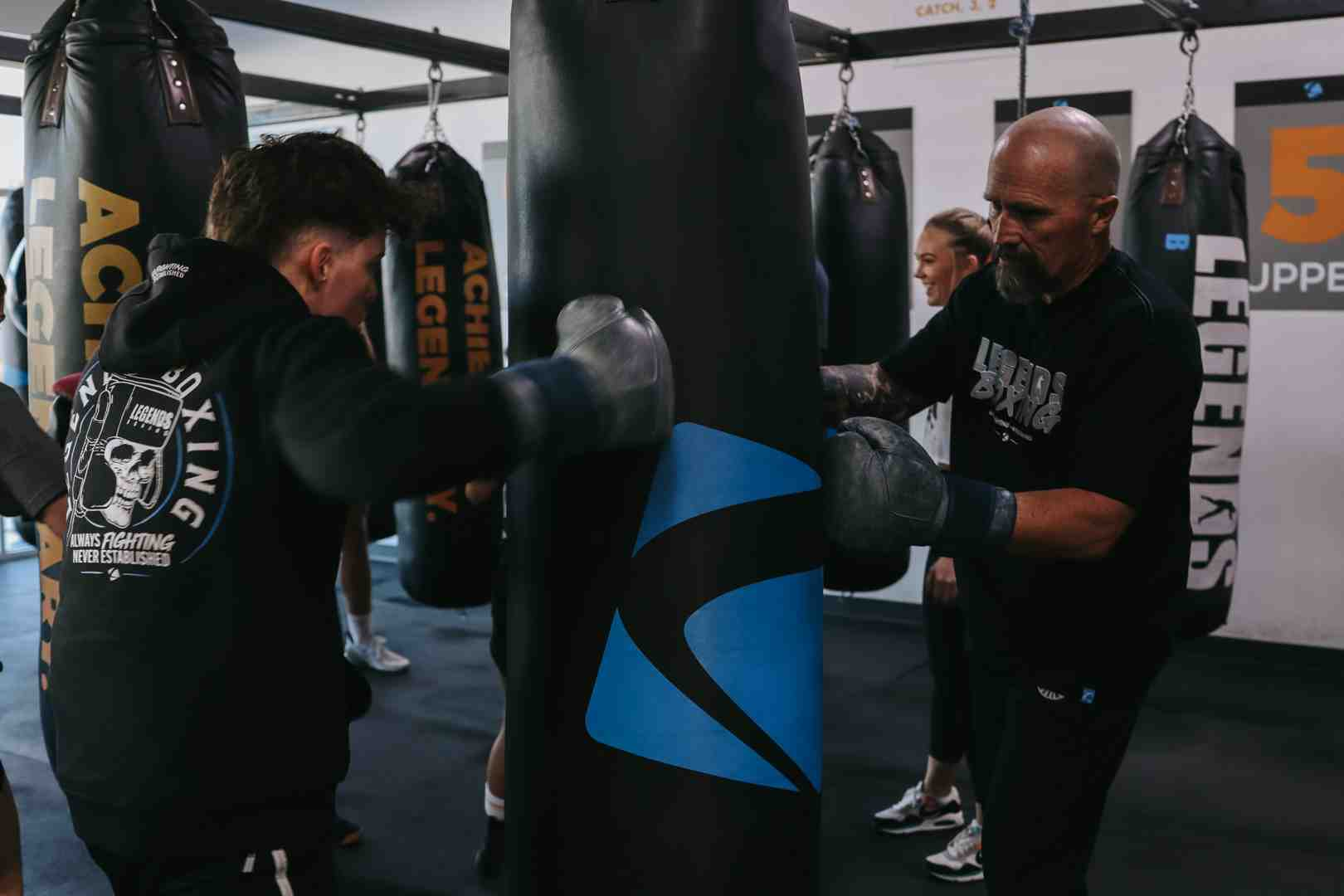 Legends Boxing coach holding a blue-and-black heavy bag steady while a student in a Legends Boxing hoodie throws a punch, full gym visible in background with rows of bags and "Achieve" posters — showing the one-on-one coaching attention that makes beginner bag work safe and effective