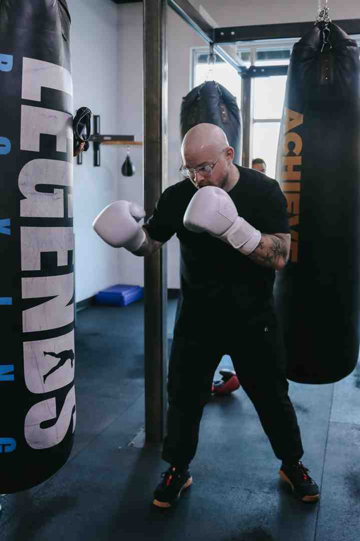 Bald boxer with glasses in all-black outfit with white Legends Boxing gloves in a proper boxing stance — left hand extended toward the LEGENDS BOXING heavy bag, ACHIEVE text visible on bag in background — boxing bag training fundamentals: stance and bag work — Legends Boxing