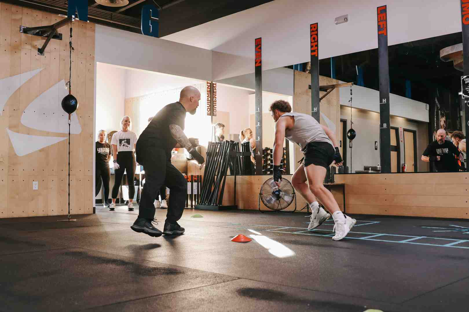 Legends Boxing coach doing one-on-one footwork drill with a member — 'RIGHT / LEFT / EVEN' signs on the wall, speed ball visible, other members observing — Salt Lake City class instruction