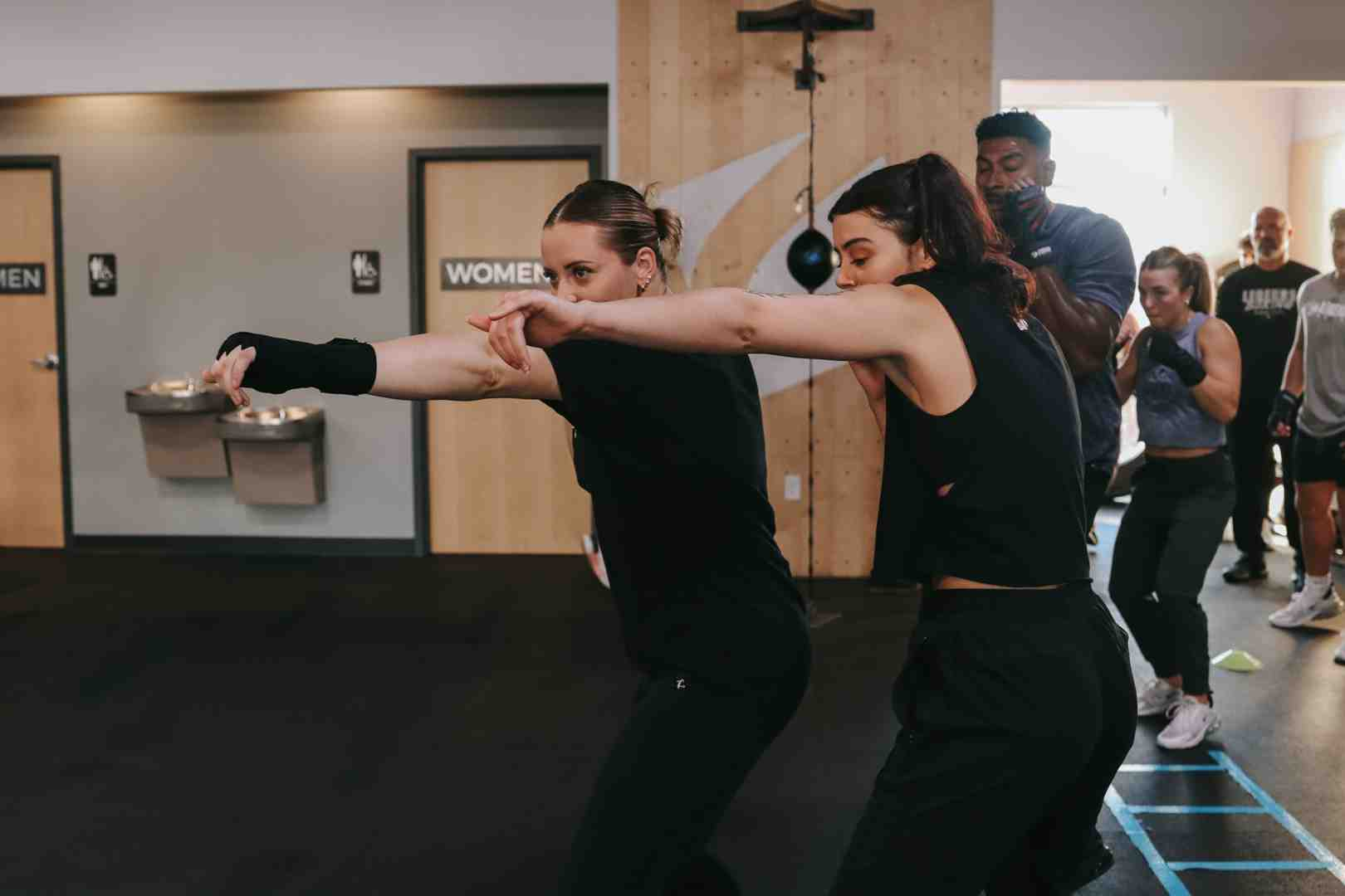 Two women training side-by-side, both throwing simultaneous crosses in a bright Legends Boxing class — supportive coaching environment, speed bag and other members visible in background — boxing for women community — Legends Boxing