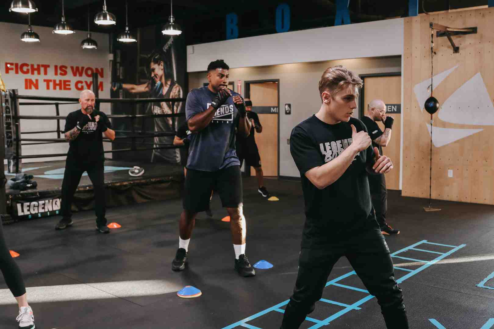 Diverse group of members in a high-energy Legends Boxing class — 'FIGHT IS WON IN THE GYM' mural on the wall, boxing ring in background, coach supervising agility drills on the floor — Bountiful, Utah