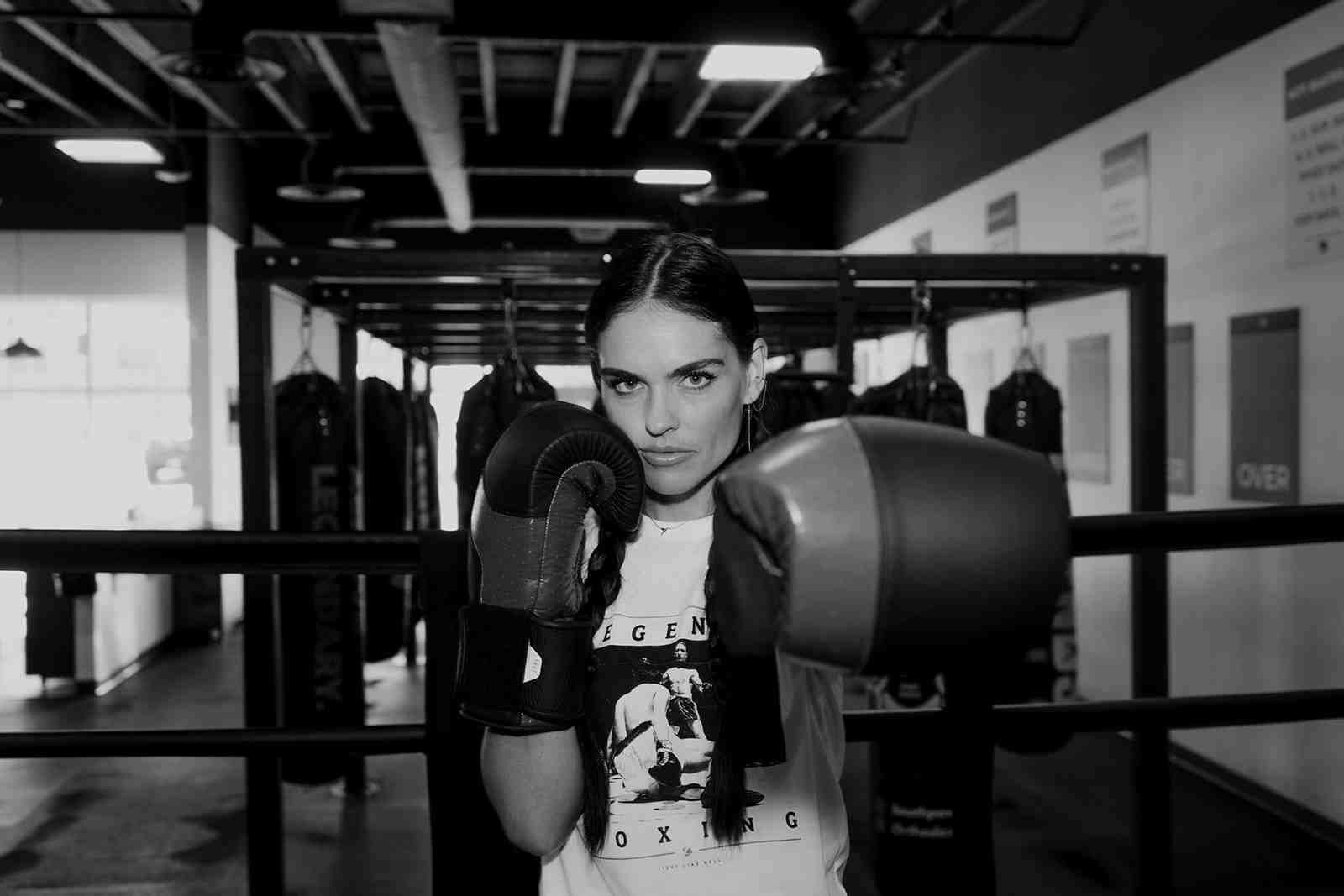 Black and white portrait of a female Legends Boxing member in guard position — gloves up, focused expression, heavy bags and boxing ring visible in the background — Bountiful gym