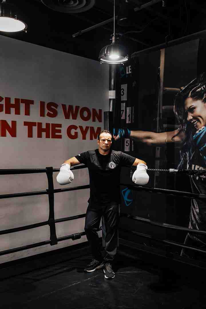 Male boxer in black outfit leaning on ring ropes with white gloves — 'THE FIGHT IS WON IN THE GYM' mural on wall behind him — Legends Boxing female athlete photo on the right — professional boxing gym Ogden Utah — Legends Boxing
