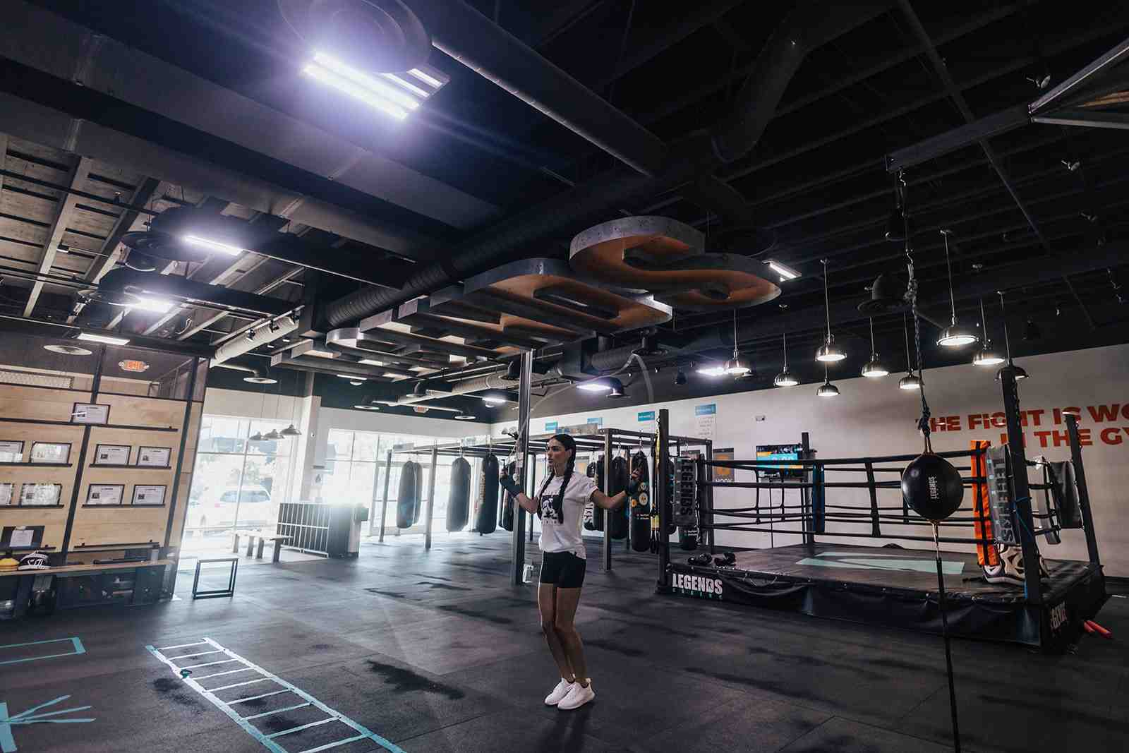 Wide interior shot of Legends Boxing Orem — woman jump-roping in foreground, full-size boxing ring in background, rows of heavy bags hanging from ceiling rig, "THE FIGHT IS WON IN THE GYM" mural on the wall — Legends Boxing