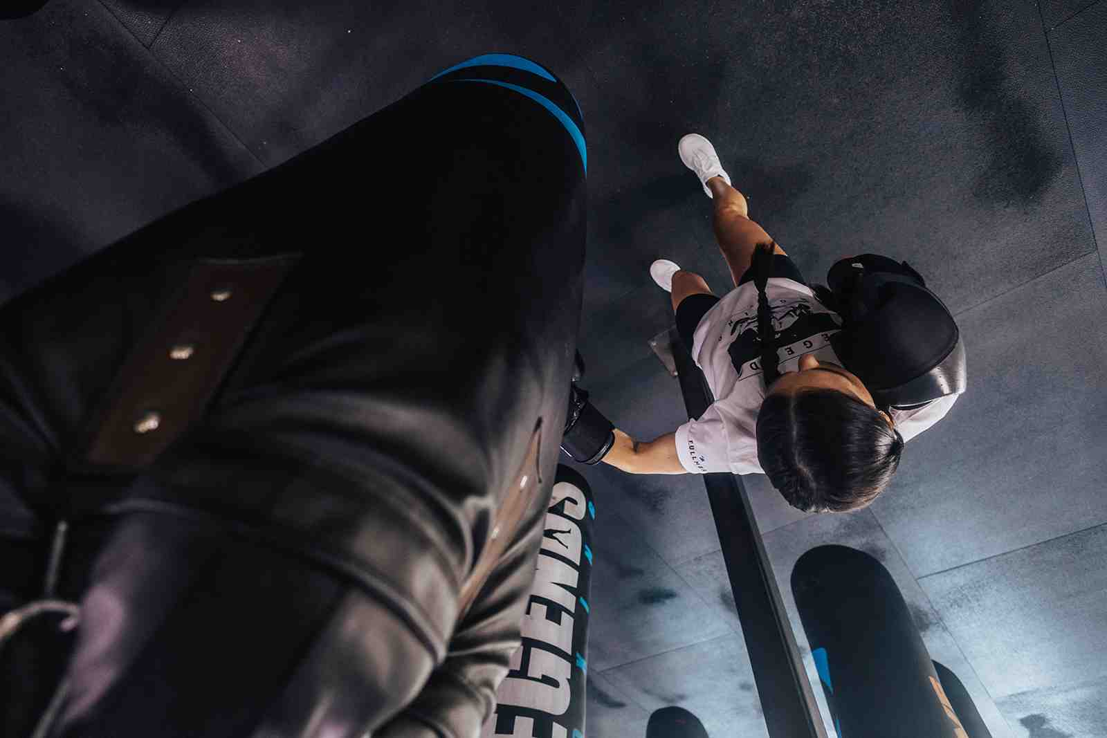 Low-angle overhead shot looking up at a female boxer in Legends Boxing gear throwing a punch at a heavy bag — dramatic ceiling perspective with other bags visible in background — boxing gym Sandy Utah — Legends Boxing