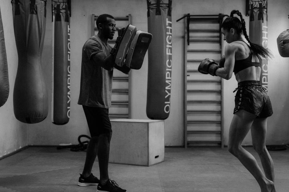 Black and white photo of a male coach holding large focus mitts for a female boxer throwing a punch — professional boxing mitt training session — heavy bags and gym equipment in background — Legends Boxing