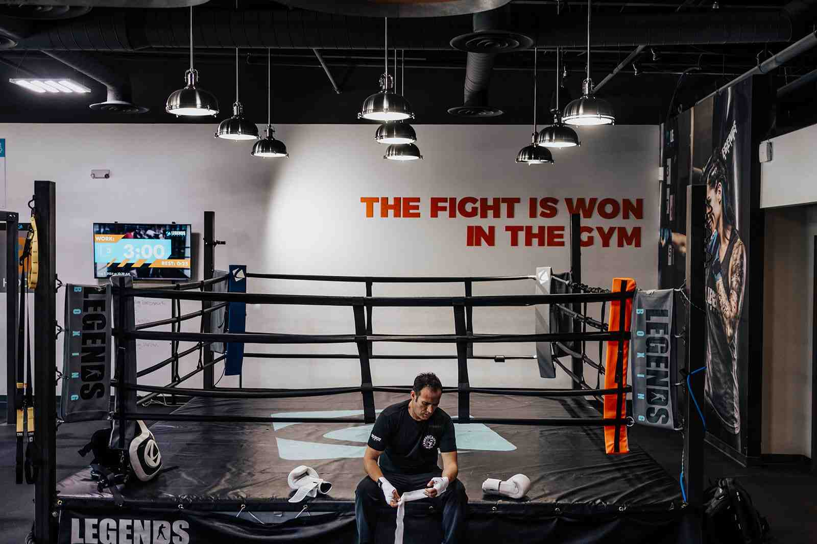 Legends Boxing ring with 'THE FIGHT IS WON IN THE GYM' mural on the wall — coach sitting on the ring apron wrapping hands, round timer showing 3:00, motivational atmosphere — boxing gym Sandy Utah — Legends Boxing