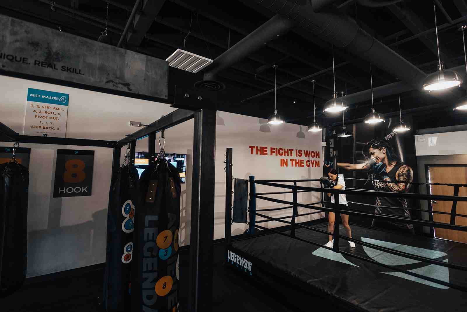 Legends Boxing gym interior — 'THE FIGHT IS WON IN THE GYM' mural in bold red, numbered target bag ('8 HOOK') and Mitt Master instruction card in foreground, woman training in the boxing ring, pendant lighting — Utah
