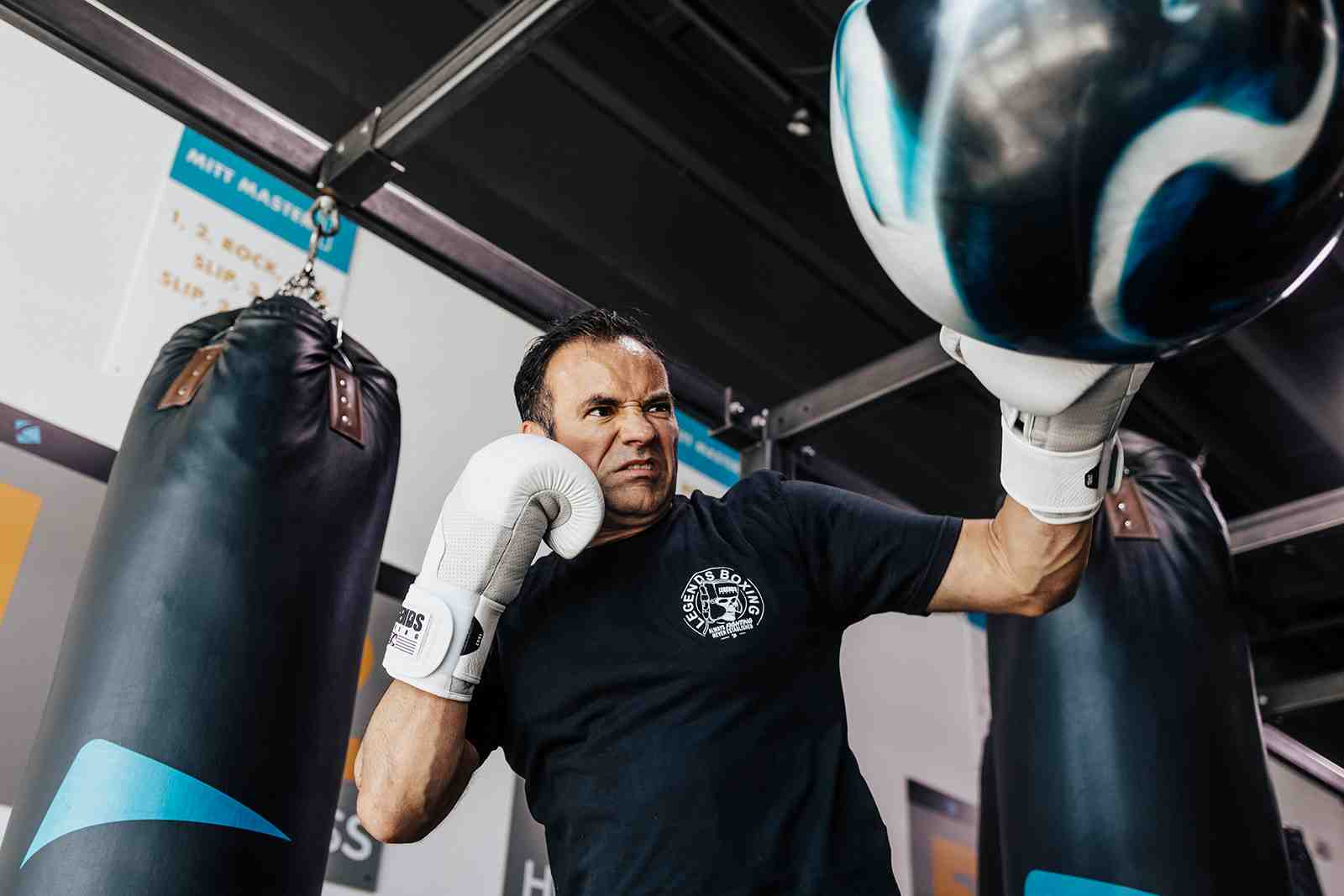 Low-angle shot of a determined man in a Legends Boxing t-shirt with white gloves, jaw set, driving an uppercut into a speed bag with his right hand while a heavy bag hangs nearby. The "MITT MASTER" coaching chart is visible on the wall above — the intense focus of generating real knockout power from the ground up