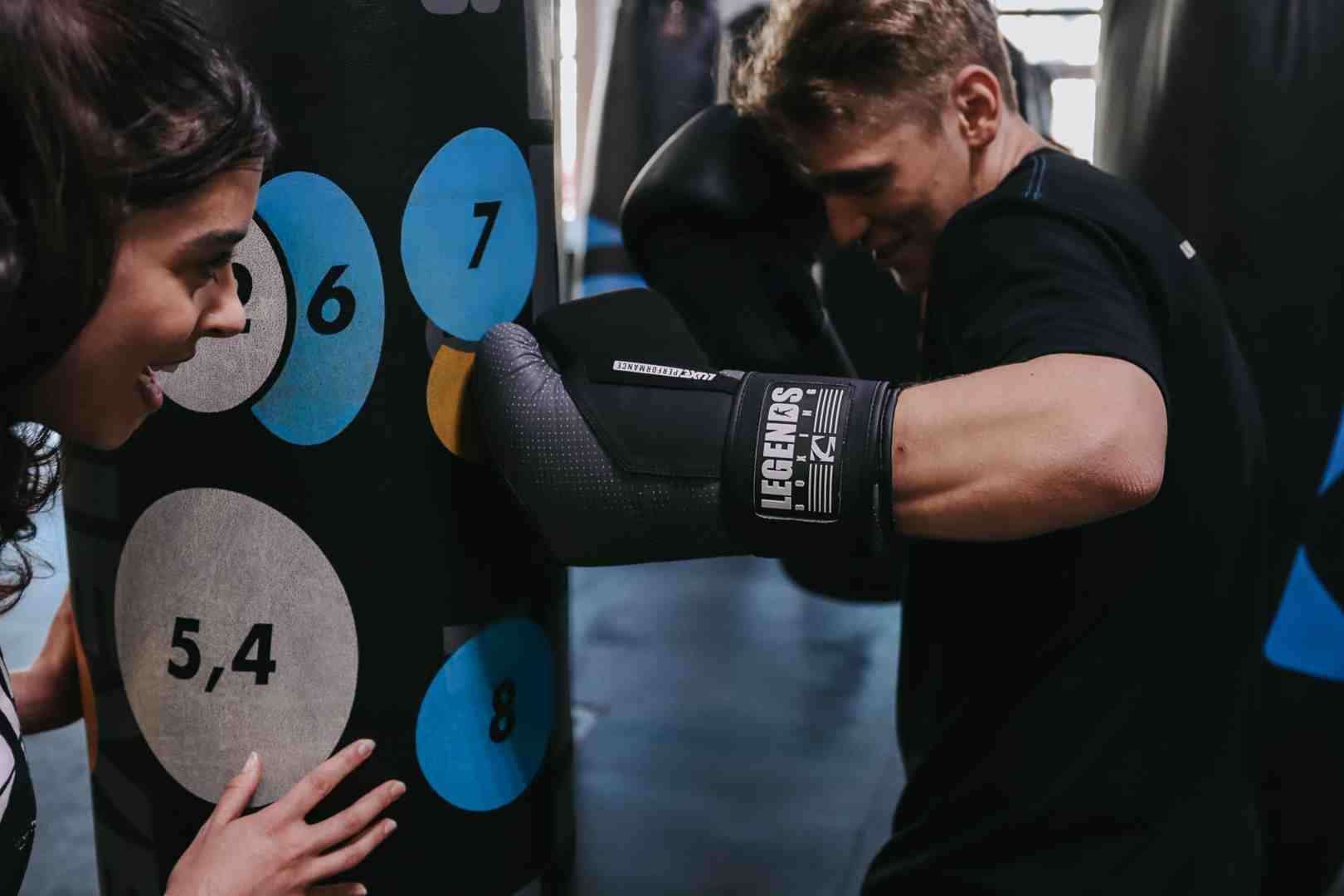 Man in Legends Boxing gloves throwing a punch at a numbered heavy bag with colored target circles (5,4 / 6 / 7 / 8), woman observing and guiding — close-up showing proper glove contact and the numbered target system that helps beginners learn punch placement