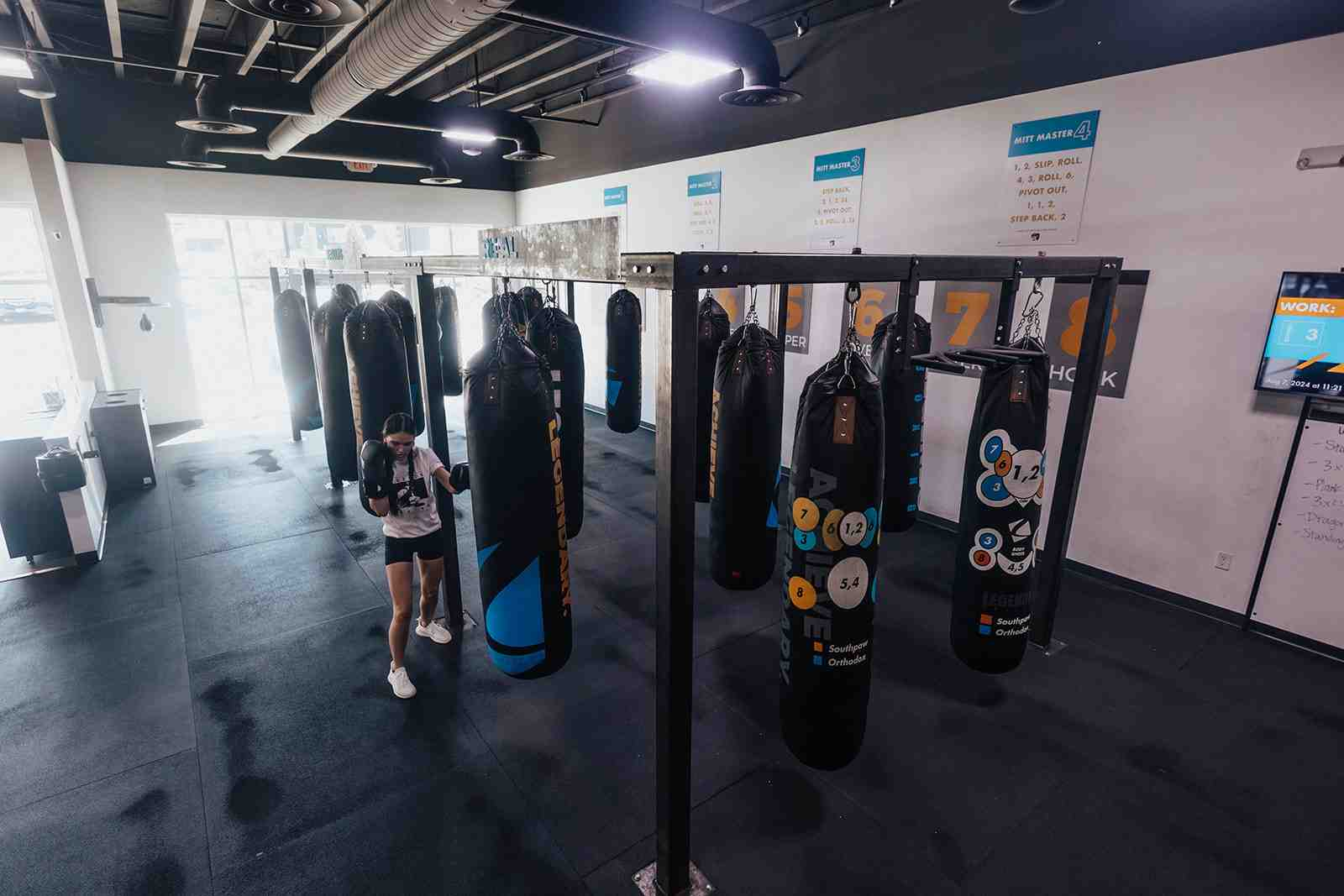 Wide-angle view of a Legends Boxing gym floor showing multiple numbered heavy bags on rack systems — Southpaw/Orthodox target circles visible, "Mitt Master" combo instruction posters on the wall, a woman training alone near the bags — showing the professional setup that a home boxing space can be modeled after
