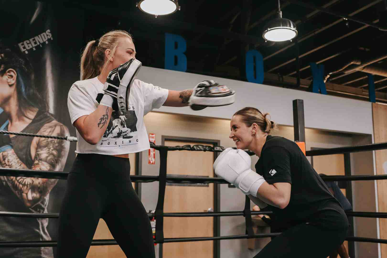 Two women practicing mitt work inside a Legends Boxing ring — one in a white Legends Boxing t-shirt extending a jab to the focus mitts, trainer in black crouching with white gloves ready to receive — "BOX" and "LEGENDS" branding on the walls — boxing moves to learn: combinations and mitt work — Legends Boxing