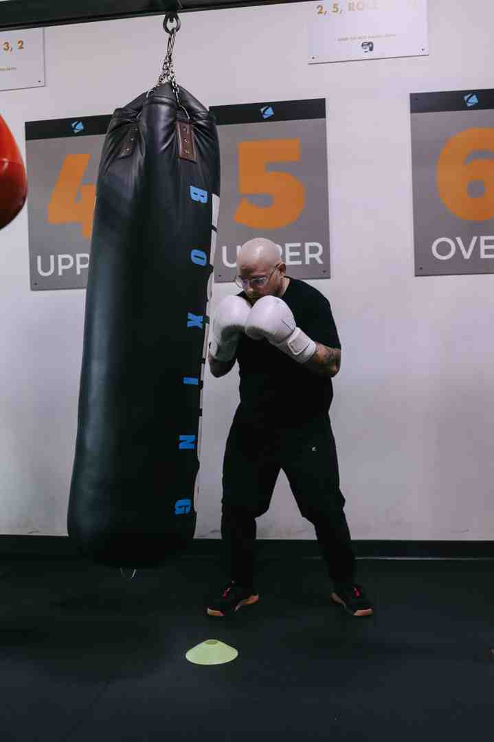 Bald male boxer in black with white gloves in a focused guard stance, working the heavy bag at Legends Boxing — numbered station posters (4 UPPER, 5 UPPER, 6 OVER) visible on the wall behind him