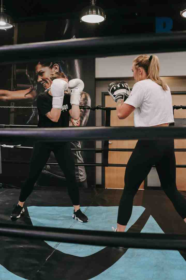 Two women boxing in the Legends Boxing ring — one in black top with white gloves, one in white top with silver mitts — viewed through the ring ropes with a Legends Boxing mural visible in the background