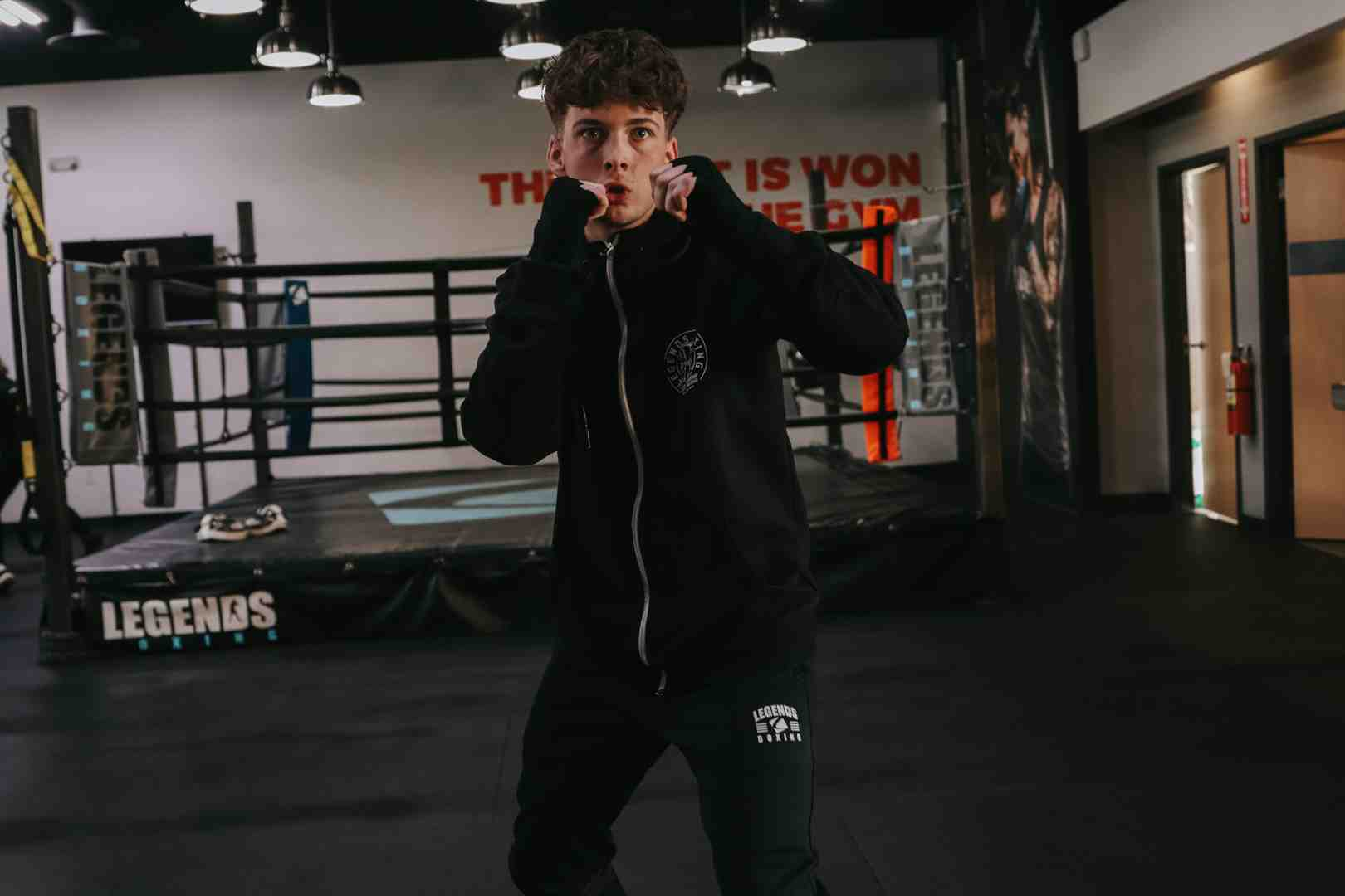 Young man in Legends Boxing hoodie and sweatpants in a tight guard position — 'THE FIGHT IS WON IN THE GYM' mural and boxing ring visible behind him — Legends Boxing gym