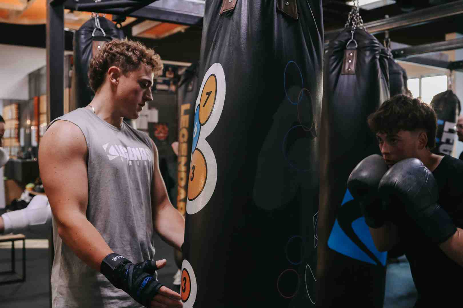 Two young men training at a Legends Boxing numbered target bag — numbers 3, 7, and 8 visible on the bag — one studying targets, one in guard position ready to punch