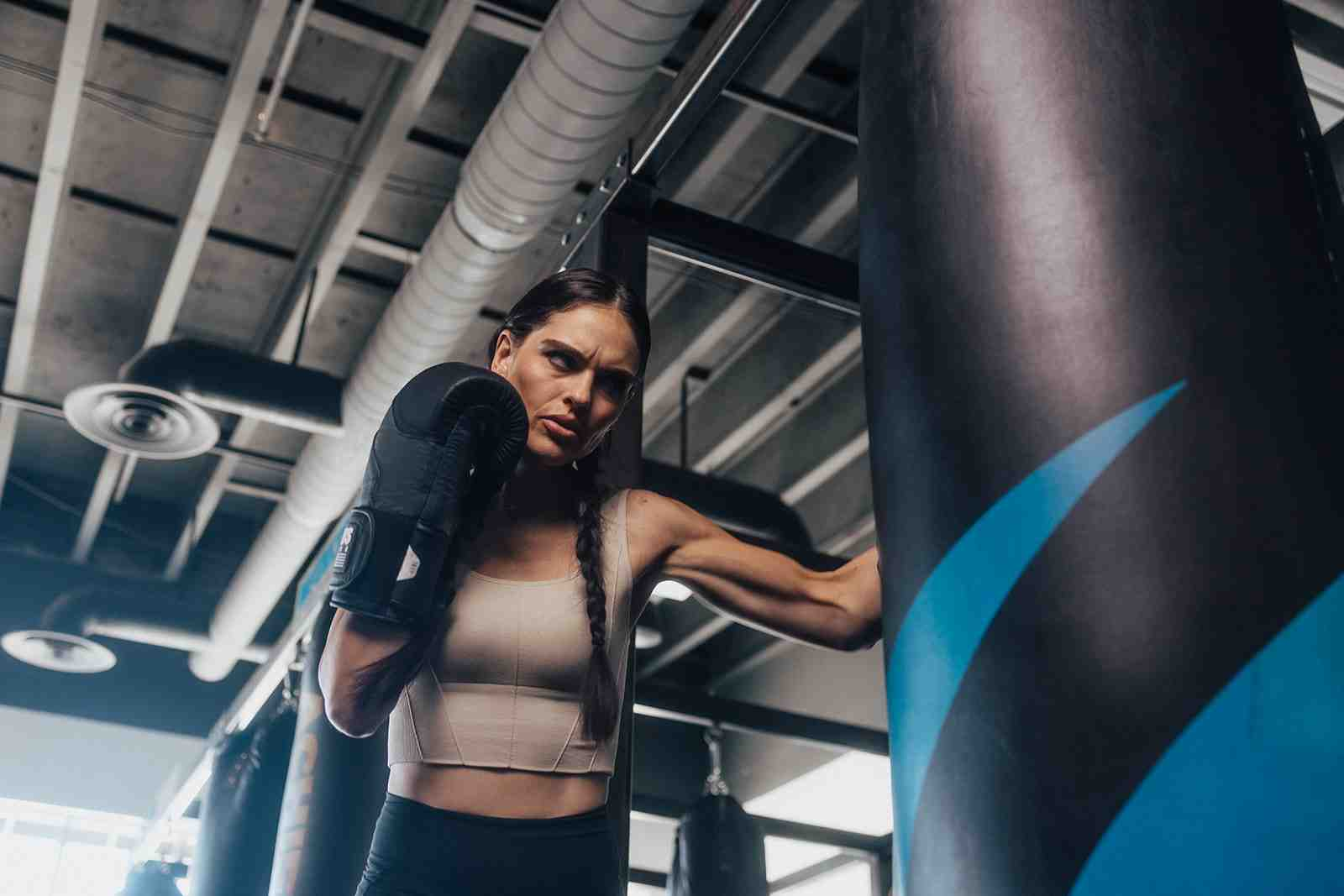 Female boxer in beige sports bra in a proper guard stance next to a Legends Boxing heavy bag — intense focused expression, side profile