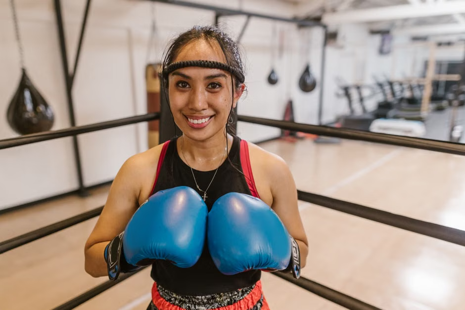 Young woman smiling confidently in large blue boxing gloves inside a boxing ring — headband, speed bags and gym equipment visible in background — fighting gyms near me: the confidence that stays with you long after class — Legends Boxing
