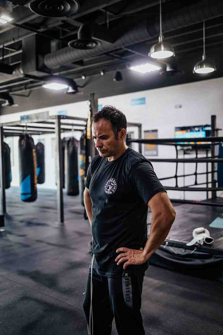 Legends Boxing coach in a black Legends Boxing shirt standing in an empty gym surrounded by rows of heavy bags and a boxing ring — surveying the training space