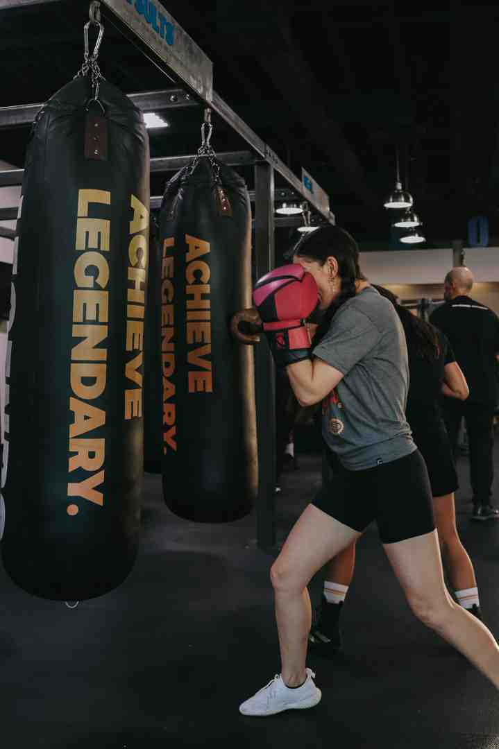 Woman in pink boxing gloves working the "Achieve Legendary" heavy bag in a Legends Boxing gym, leaning into a body shot with proper form