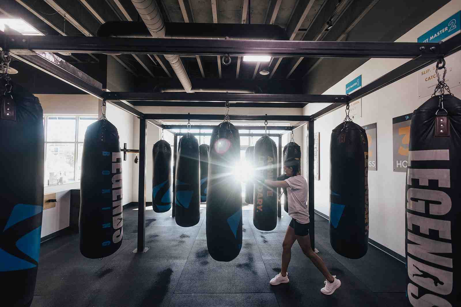 Woman throwing a punch at a heavy bag at the end of a long corridor of Legends Boxing heavy bags, dramatic backlighting streaming through windows behind her, "Mitt Master" instruction posters visible on the wall — showing the focused intensity that defines heavy bag interval training sessions