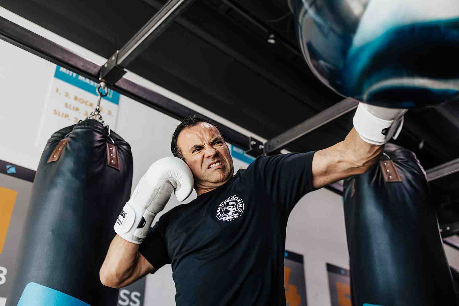 Legends Boxing coach in a Legends Boxing t-shirt driving a close-range punch into a heavy bag from a dramatic low angle, a second bag visible beside him — showing the full-body power transfer and intense focus that separate power-building drills from casual bag work