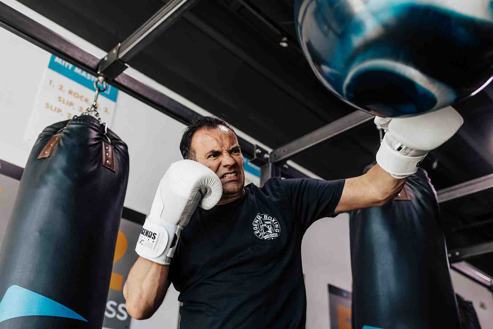 Intense man in Legends Boxing shirt throwing a powerful punch with white gloves between two heavy bags — focused expression showing drive and commitment — power shot training at Legends Boxing