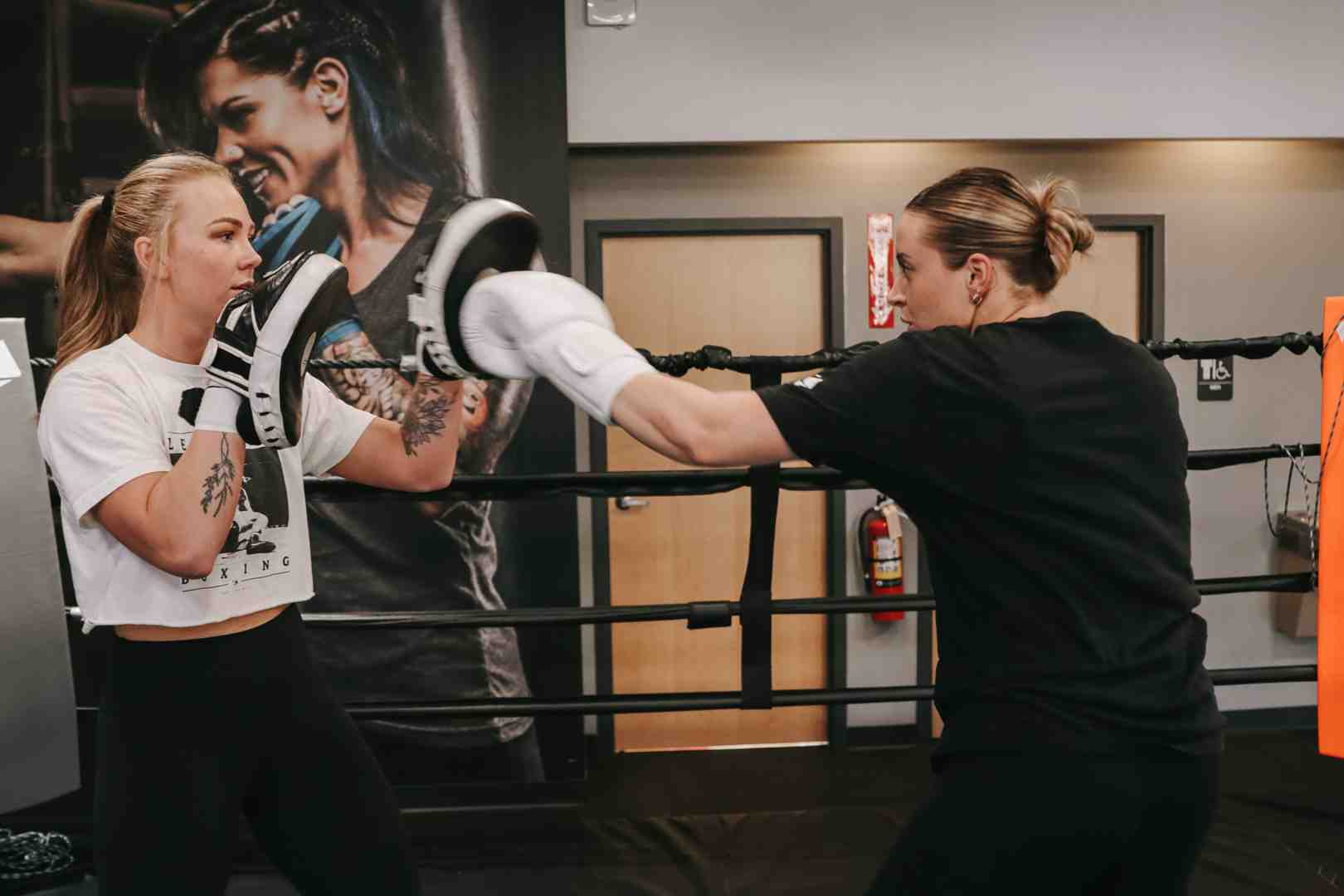 Two women doing focus mitt work in a Legends Boxing ring — one in white Legends tee holding mitts, one in black throwing a jab — large Legends athlete mural on wall behind them — Legends Boxing
