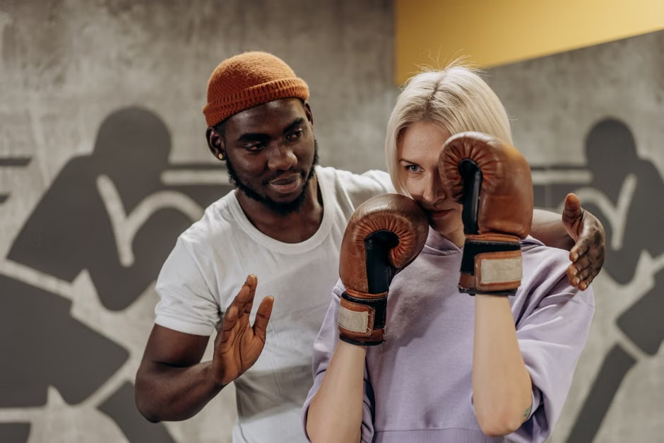 Male boxing coach in orange beanie adjusting a female boxer's guard position — woman in lavender top with tan brown leather gloves holding defensive stance — boxing murals on wall behind them — Legends Boxing