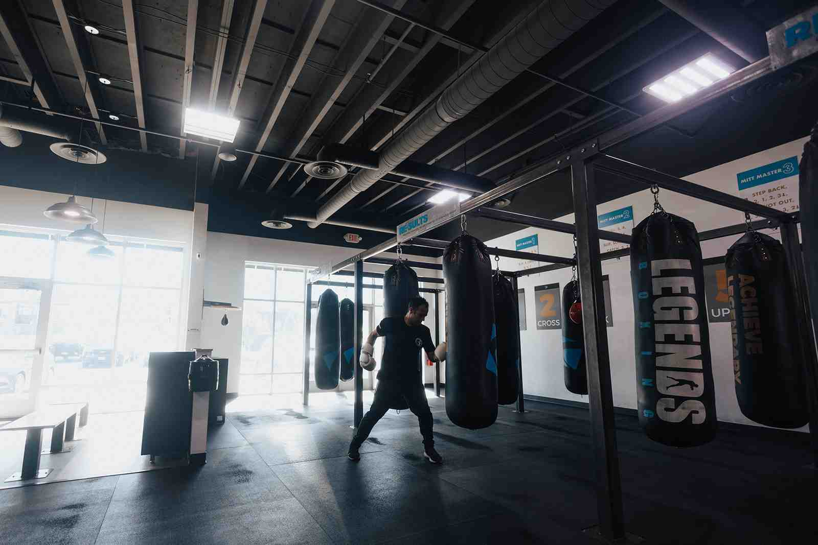 Wide-angle interior of a Legends Boxing gym — solo boxer working heavy bags in the center, row of 'LEGENDS' branded bags extending to the back, 'ACHIEVE' and 'CROSS' instruction signs on the walls, natural light from large windows — Legends Boxing Utah