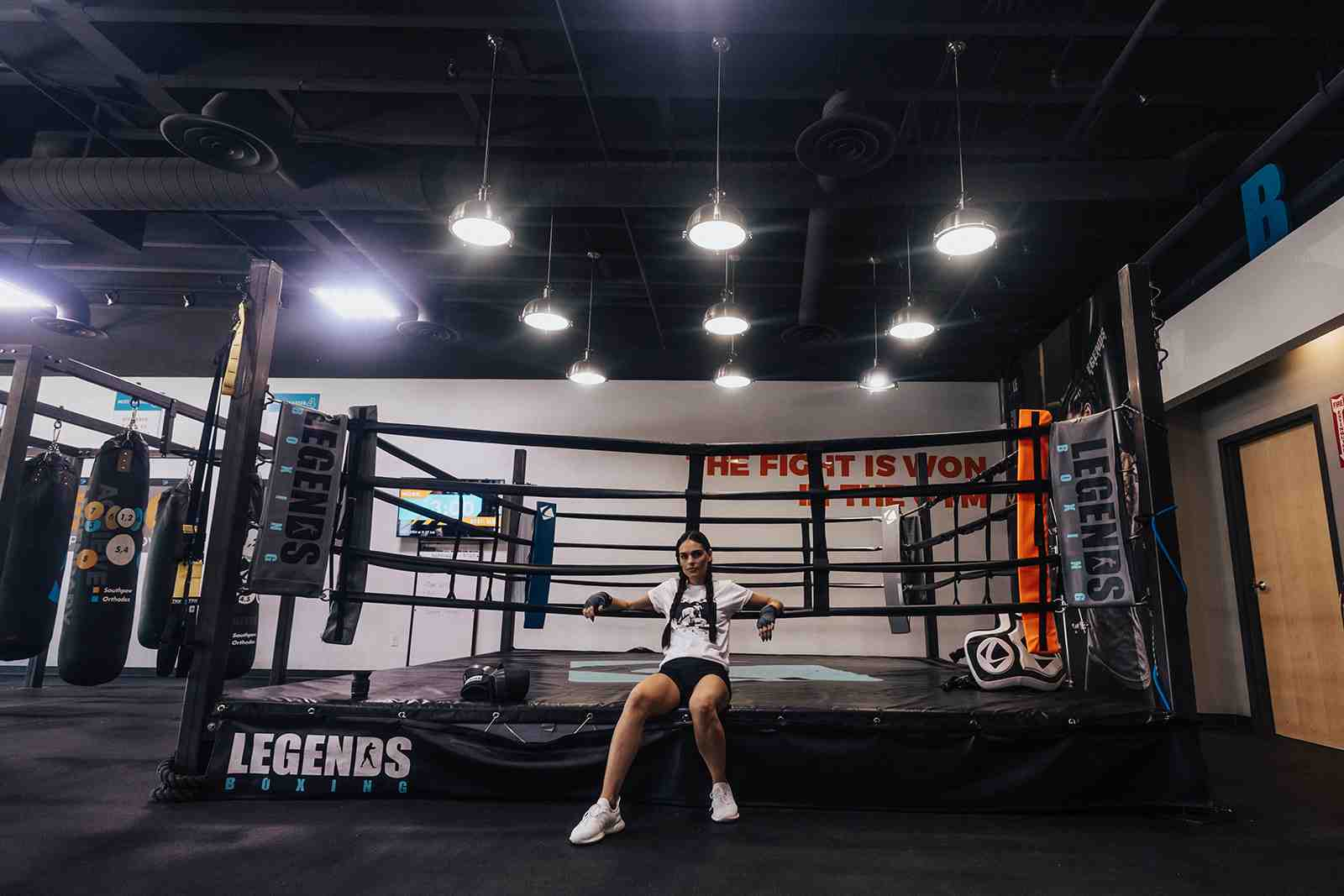 Clean, modern Legends Boxing gym interior — woman sitting on the edge of a full-size boxing ring, 'THE FIGHT IS WON IN THE GYM' painted on the wall, heavy bags on the left, pendant lighting — Salt Lake City area
