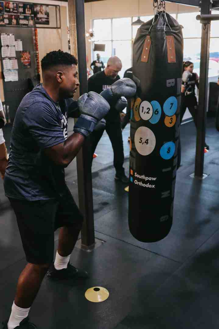 Man with proper form hitting a numbered target heavy bag (Southpaw/Orthodox color-coded circles) at Legends Boxing — coach and other members training in the background, Salt Lake City